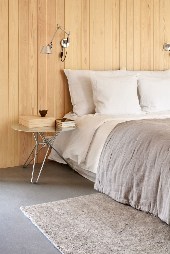 Roots Living pellavalakanoilla pedattu sänky makuuhuoneessa, jossa on paneeliseinät ja betonilattia. Lattialla pellavamatto.