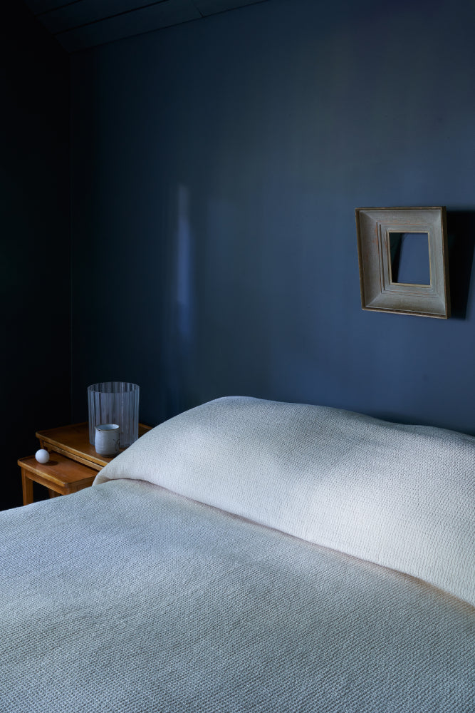 Luonnonvalkoisella Polkupäiväpeitolla pedattu sänky tummansinistä seinää vasten. Seinällä kehykset.
