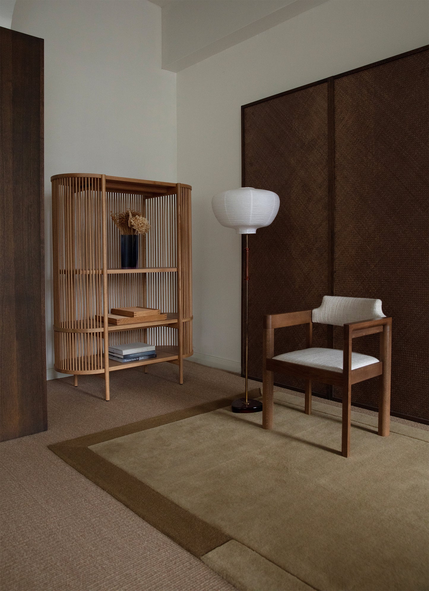 Khakin sävyinen Geo One -villamatto puisen verhoillun tuolin alla. Tuolin vieressä jalkavalaisin. Maton vieressä puinen hylly. Kalusteita ympäröi valkoiset seinät sekä pähkinän sävyisen puupaneelit.