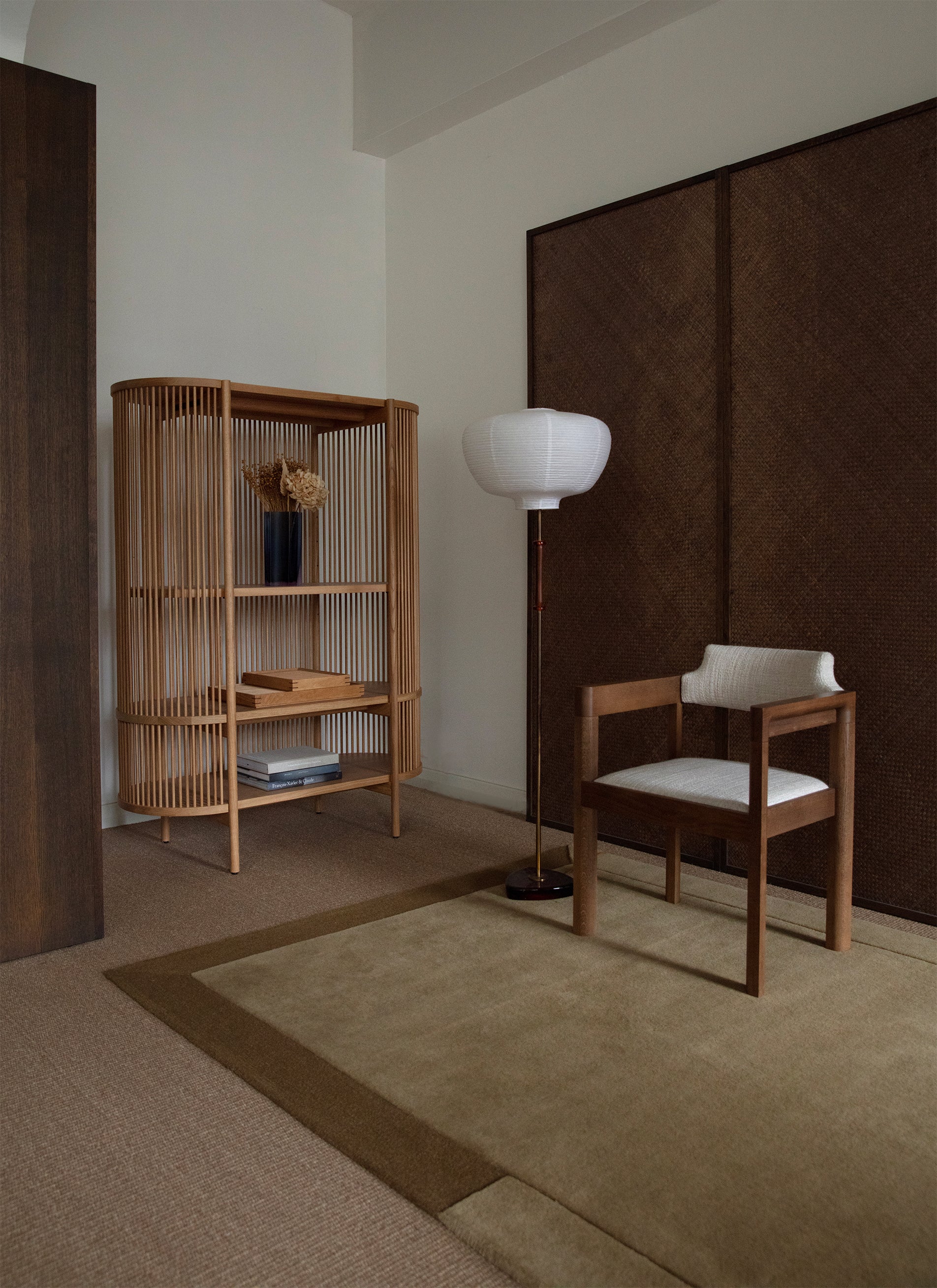 Khakin sävyinen Geo One -villamatto puisen verhoillun tuolin alla. Tuolin vieressä jalkavalaisin. Maton vieressä puinen hylly. Kalusteita ympäröi valkoiset seinät sekä pähkinän sävyisen puupaneelit.