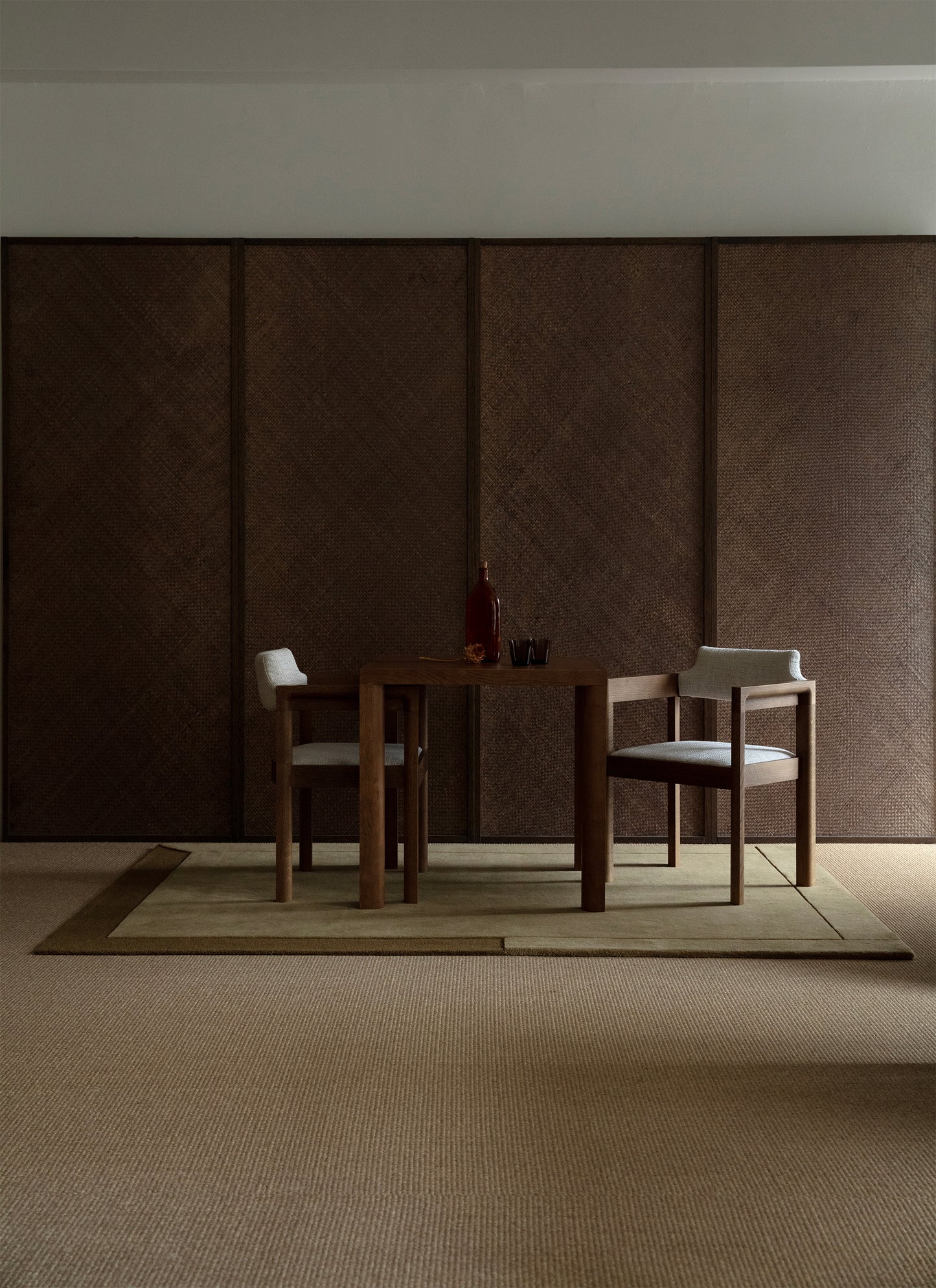 Puinen pöytäryhmä kuvattuna pähkinän sävyistä seinää vasten. Pöytäryhmän alla khakin sävyinen Geo One -villamatto.